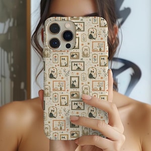 May include: A beige phone case featuring a repeating pattern of black and white cats in gold frames. The cats are surrounded by small floral and geometric designs.