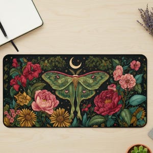 May include: A rectangular black desk mat featuring a green moth with a crescent moon above it, surrounded by colorful flowers. The flowers include red, pink, and yellow blooms with green foliage. The background is a dark starry night.