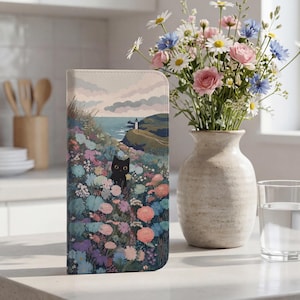 May include: A wallet case featuring a coastal landscape with a black cat peeking through colorful flowers. The design includes a lighthouse, ocean, and sky. A vase of flowers and a glass of water are in the background.