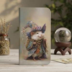 Puede incluir: Una funda rectangular para teléfono con la ilustración impresa de un ratón pequeño, blanco y esponjoso que lleva un sombrero puntiagudo azul y morado y una capa a juego, sosteniendo un ramo de flores secas sobre un fondo claro y suave.
