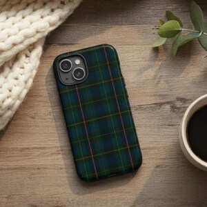 May include: A smartphone case featuring a green and navy tartan design. The case has a black camera module and is on a wooden surface. A white knitted scarf and a cup of coffee are also visible.