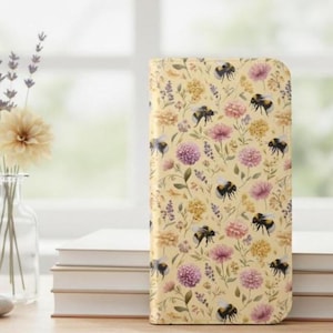 May include: A floral patterned phone case featuring bees, purple and pink flowers, and green leaves on a light yellow background. The case is standing upright on a stack of white books.