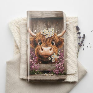 Puede incluir: Una funda de teléfono ilustrada con una vaca de las Tierras Altas marrón y blanca con una corona de margaritas, mirando a través de una valla de madera. La vaca está rodeada de flores moradas y pequeños corazones blancos. La funda descansa sobre una manta de punto beige.