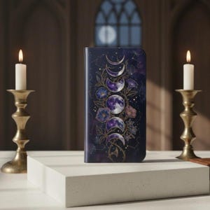 Op de afbeelding: Een donkerpaarse telefoonhoes met een hemels ontwerp met maanfasen, bloemen en sterren. De hoes staat tegen een achtergrond met brandende kaarsen in gouden kandelaars.