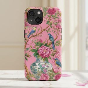May include: Pink phone case featuring a floral and bird design. The case has a pink background with a pattern of pink flowers, green leaves, and blue birds perched on branches. A decorative vase with flowers is in the center.