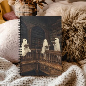 May include: A spiral-bound notebook featuring a spooky illustration of three ghosts reading books in a library. The artwork is detailed with a vintage aesthetic, showcasing wooden bookshelves and a balustrade. The ghosts are depicted in white sheets.