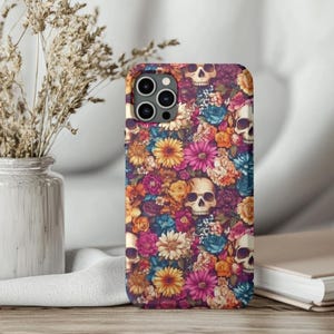 May include: A phone case featuring a vibrant floral and skull pattern. The design incorporates a mix of colorful flowers in shades of pink, orange, yellow, and blue, set against a dark background. Several white skulls are interspersed throughout the floral arrangement.