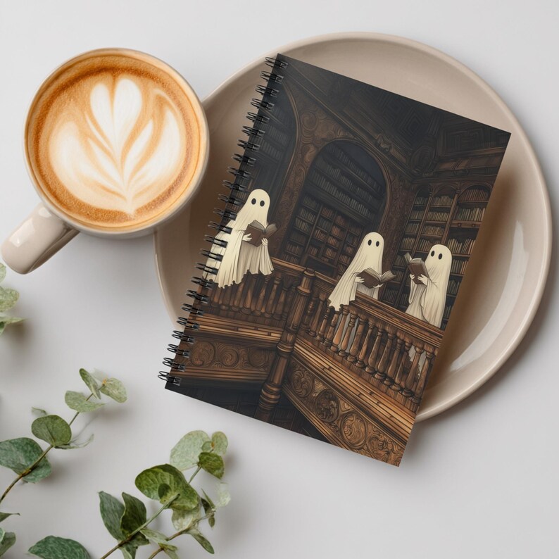 May include: Spiral-bound notebook featuring a spooky illustration of three ghosts reading books in a library. The cover art is set against a backdrop of a vintage wooden staircase and bookshelves. A cup of coffee with latte art sits next to the notebook.