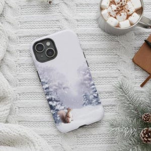 Puede incluir: Funda de teléfono blanca con un diseño de bosque invernal y una ardilla. La funda tiene un módulo de cámara negro y botones laterales negros. El fondo incluye una taza de chocolate caliente con malvaviscos, un cuaderno y piñas.
