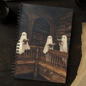 May include: Spiral-bound notebook featuring a spooky illustration of three ghosts reading books in a library. The artwork is in shades of brown and beige, with a detailed depiction of bookshelves and a wooden staircase. The cover art has a vintage, gothic aesthetic.