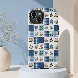 Könnte beinhalten: Eine Smartphone-Hülle mit einem Gittermuster aus abwechselnden Quadraten. Das Design umfasst blau-weiß karierte Vichy-Muster, einfarbige dunkelblaue Quadrate und weiße Quadrate, die mit verschiedenen Blumenillustrationen wie rosa Rosen, blauen Hortensien und Gänseblümchen verziert sind.