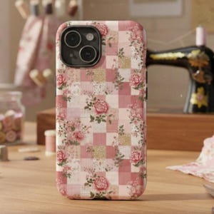 Könnte beinhalten: Eine Handyhülle mit floralem Muster und Patchwork-Design. Die Hülle zeigt eine Mischung aus rosa und weißen Quadraten, die mit rosa Rosen und grünen Blättern verziert sind. Die Handyhülle liegt auf einer Holzoberfläche.