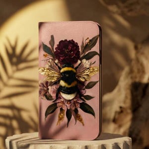 May include: A pink phone case featuring a detailed illustration of a bumblebee with golden wings, surrounded by flowers and foliage. The design includes a dark red flower at the top and pink flowers at the bottom, creating a nature-inspired theme.