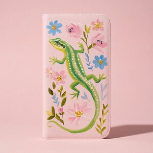 Pode incluir: Uma carteira rosa claro com um desenho pintado à mão de um lagarto verde com manchas brancas, rodeado por flores rosa e azuis e folhas verdes. A carteira tem uma textura suave e uma forma retangular.