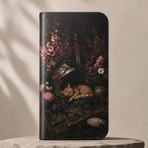 May include: A black rectangular wallet featuring a printed scene of a sleeping orange tabby cat nestled beside a metallic helmet, a sword, and a stone, all surrounded by dark green foliage and clusters of pink and white flowers.