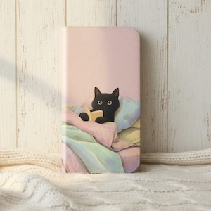 Op de afbeelding: Roze telefoonhoesje met een zwarte kat die in bed leest, onder een pastelkleurige deken en kussen. De hoes heeft een gladde afwerking en een magnetische sluiting.