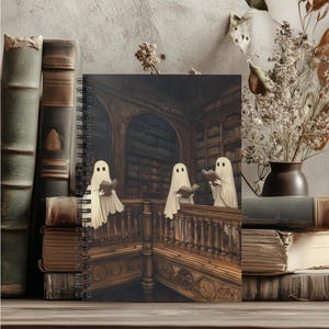 May include: A spiral-bound notebook featuring a spooky illustration of three ghosts reading books in a library. The cover art is set against a backdrop of antique books and a wooden surface, evoking a vintage aesthetic.