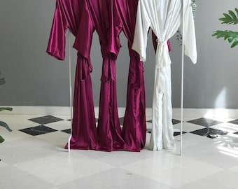 以下が含まれることがあります： 白い金属製のラックに掛けられた4着の長袖ローブ。3着は光沢のあるサテン仕上げの深いマゼンタ色で、1着は同じく光沢のあるサテン仕上げの白色です。各ローブにはウエストを結ぶための共布のベルトが付いており、袖はゆったりとしたデザインです。
