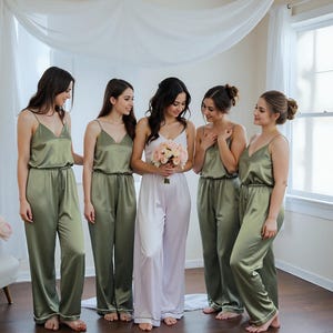 A bride in white satin bridal pajamas holds a bouquet, surrounded by her bridesmaids in matching olive green camisole pajama sets. The luxurious satin outfits feature full-length pants and a relaxed fit, perfect for getting-ready moments and bridal party gifts. A chic example of personalized women's clothing for weddings.