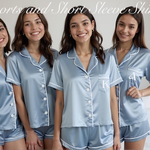 Four women stand side by side in matching pastel blue satin pajama sets, each featuring short-sleeve button-down shirts with white piping and coordinating shorts. Two of the shirts display white personalized initials, "K" and "A," on the front pocket. They are all smiling in a bright bedroom setting. The image highlights the option of monogrammed sleepwear for bridal parties or group gifting. Text at the top reads: "Shorts and Short Sleeve Shirts."