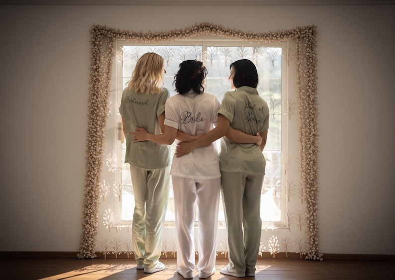 Women dressed in elegant satin pajama sets in Olive and white, each personalized with embroidered names or initials. They pose together in a cozy, well-lit bedroom setting, showcasing the luxurious and stylish design, ideal for special occasions.
