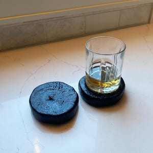 Puede incluir: Un vaso transparente con líquido ámbar descansa sobre un posavasos negro. Hay otro posavasos negro similar al lado. Los posavasos parecen estar hechos de un material oscuro y texturizado, posiblemente madera.