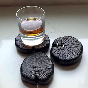 May include: Three circular coasters made from charred, black-stained wood slices featuring visible grain patterns and metal staples across the centre cracks. A clear glass containing an amber-coloured liquid rests on one of the wooden coasters.