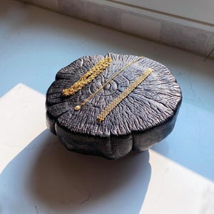 Op de afbeelding: Drie goudkleurige armbanden op een donker, verkoold cirkelvormig houten blok. Eén armband heeft een dikke gevlochten ketting, een andere is een dunne delicate ketting met een kleine parel en de derde is een brede armband met gaasstructuur.