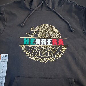 Pode incluir: Camisola de capuz preta com um gráfico dourado e verde do brasão de armas mexicano e a palavra "HERRERA" impressa no centro.