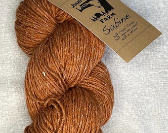 May include: A skein of brown yarn labeled "Sabine" from Juniper Moon Farm. The tag indicates the yarn is a blend of royal llama, merino wool, and cotton. The yarn is a warm, earthy tone, perfect for knitting or crocheting projects.