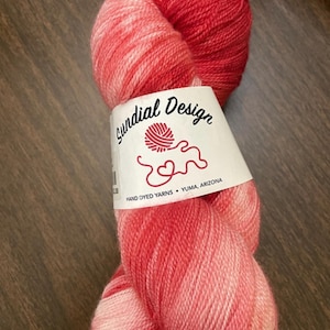 May include: A ball of hand-dyed yarn in red and pink hues. The yarn displays a gradient effect, shifting from a deep red to a lighter pink. A label reads "Sundial Design, Hand Dyed Yarns, Yuma, Arizona."