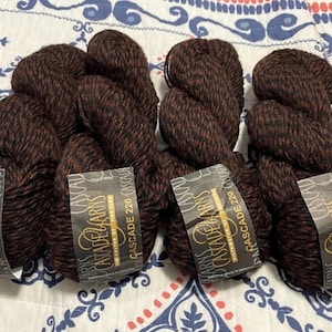 May include: Several skeins of Cascade 220 yarn in a dark brown color with red flecks. Each skein has a label that reads "Cascade 220". The yarn is arranged on a patterned fabric.