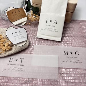 Könnte beinhalten: Hochzeitsgeschenke aus weißem Papier mit aufgedrucktem Text. Der Text enthält Initialen, ein Herzsymbol und ein Datum. Die Gastgeschenke werden auf einer rosa Oberfläche mit Trockenblumen und einer Holzkiste im Hintergrund präsentiert.