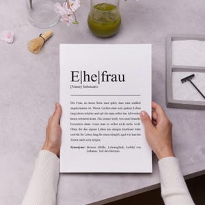 Stilvolle personalisierte Wandkunst mit Definition von Ehefrau. Hände halten ein elegantes Poster. Perfektes Geschenk für Frauen. Geschmackvolle Hintergrundelemente wie Matcha und Blumen für eine ästhetische Präsentation.