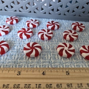 May include: A collection of red and white peppermint candies arranged on a light blue textured surface. A wooden ruler with inch markings is in the foreground. A gray plastic basket is in the background.