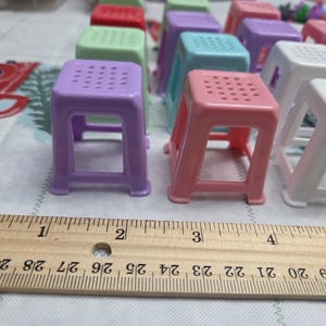 May include: A collection of small, colorful plastic stools in various shades including purple, pink, and white. Each stool has a square seat with a grid of holes and four legs. A wooden ruler is placed in front of the stools.