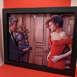 Puede incluir: Una pieza de arte pop-up 3D enmarcada que presenta a un hombre con esmoquin sosteniendo una caja azul y una mujer con un vestido rojo y guantes blancos. El fondo es un interior rosa y marrón con una puerta.