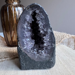 May include: A large, upright amethyst geode featuring a dark, rough exterior and a hollow centre filled with deep purple, translucent, pointed crystal formations. The geode has a jagged, irregular opening that reveals the crystalline structure.