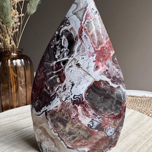 Peut inclure: Une grande sculpture en pierre en forme de goutte d'eau avec une surface polie. La pierre présente un motif complexe de tourbillons et de veines rouges, blancs, gris et noirs. La sculpture est exposée sur une surface en bois clair.