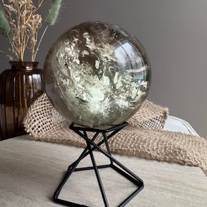 May include: A large, translucent, smoky-toned quartz sphere with visible internal fractures and inclusions, resting securely on a black, geometric, four-legged metal wire stand that features a square base and a cross-braced support structure.