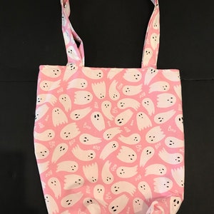May include: A pink tote bag with a white ghost pattern. The ghosts have black eyes and mouths and the words "Boo" are scattered throughout the pattern.