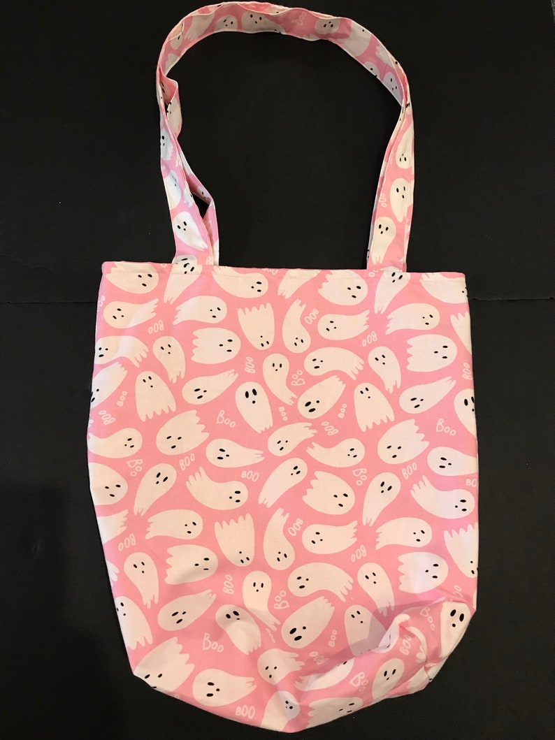 May include: A pink tote bag with a white ghost pattern. The ghosts have black eyes and mouths and the words "Boo" are scattered throughout the pattern.