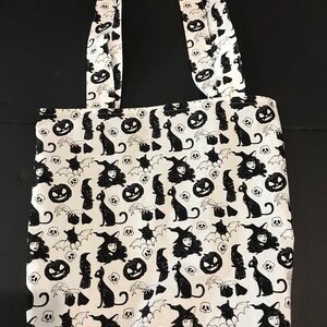 May include: A white tote bag with black Halloween-themed print. The print features witches, cats, bats, pumpkins, and skulls.