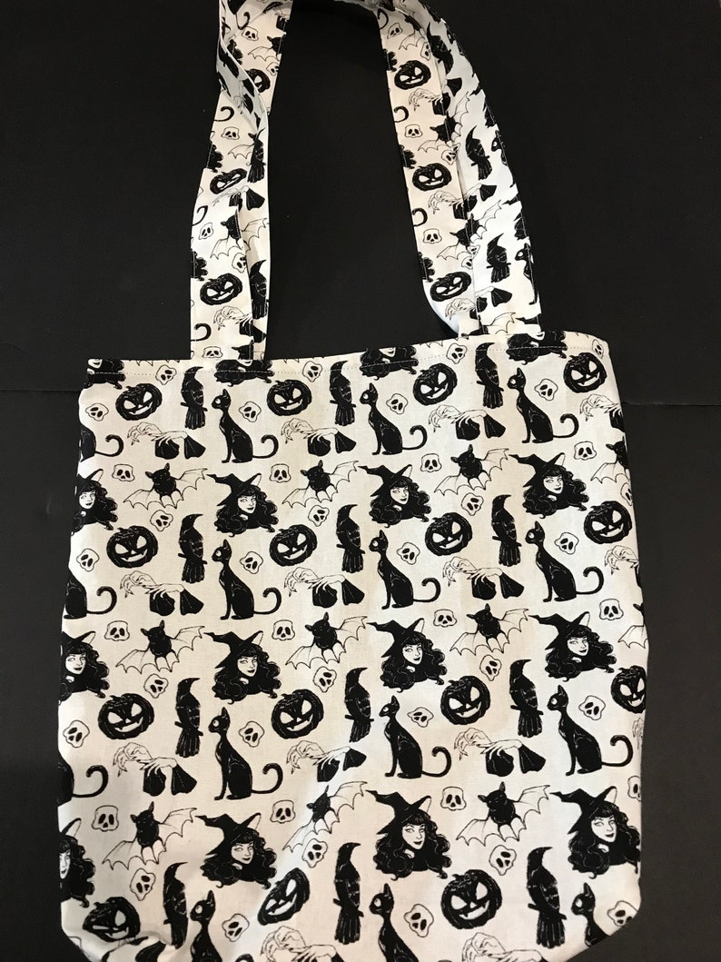 May include: A white tote bag with black Halloween-themed print. The print features witches, cats, bats, pumpkins, and skulls.