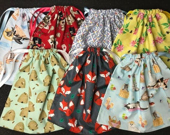 May include: A collection of eight drawstring bags made of fabric with various patterns. The patterns include bees and honeycombs, foxes, cats, flowers, and playing cards. The bags are all different colors, including blue, green, red, yellow, and gray.