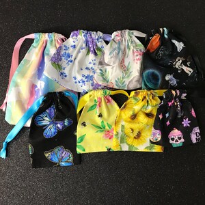 May include: A selection of small drawstring bags with diverse patterns. Designs include unicorns, floral prints, butterflies, bees, sunflowers, and space themes. Each bag has a matching ribbon drawstring closure.