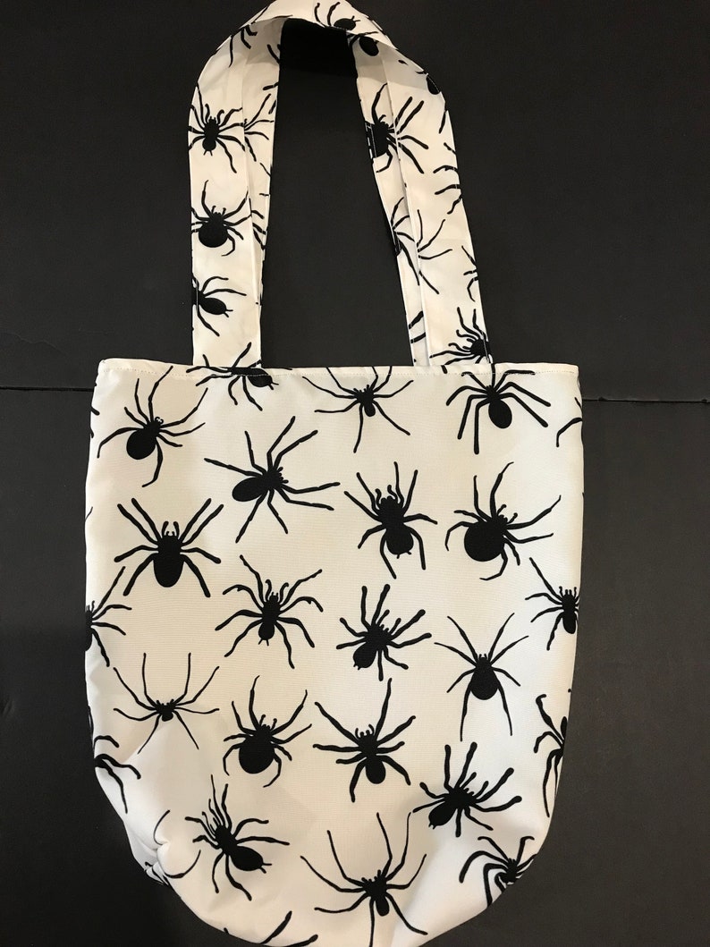 May include: White tote bag with black spider silhouettes printed all over. The bag has two black straps.