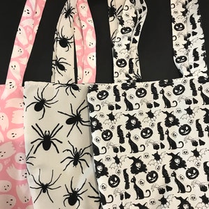 May include: Three Halloween-themed tote bags. The first bag is pink with white ghost silhouettes and the text "Boo". The second bag is white with black spider silhouettes. The third bag is white with black silhouettes of cats, bats, pumpkins, and witches.