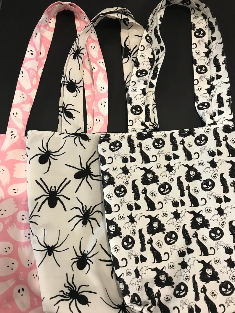 May include: Three Halloween-themed tote bags. The first bag is pink with white ghost silhouettes and the text "Boo". The second bag is white with black spider silhouettes. The third bag is white with black silhouettes of cats, bats, pumpkins, and witches.