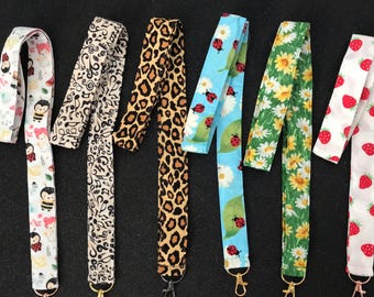 May include: Six fabric lanyards with metal lobster clasps. Patterns include cartoon bees, black musical notes on beige, brown leopard print, ladybugs on light blue, yellow sunflowers on green, and red strawberries on a white background.